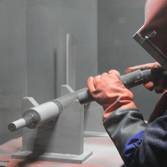 A worker in orange gloves and protective gear machines a metal rod on a lathe.